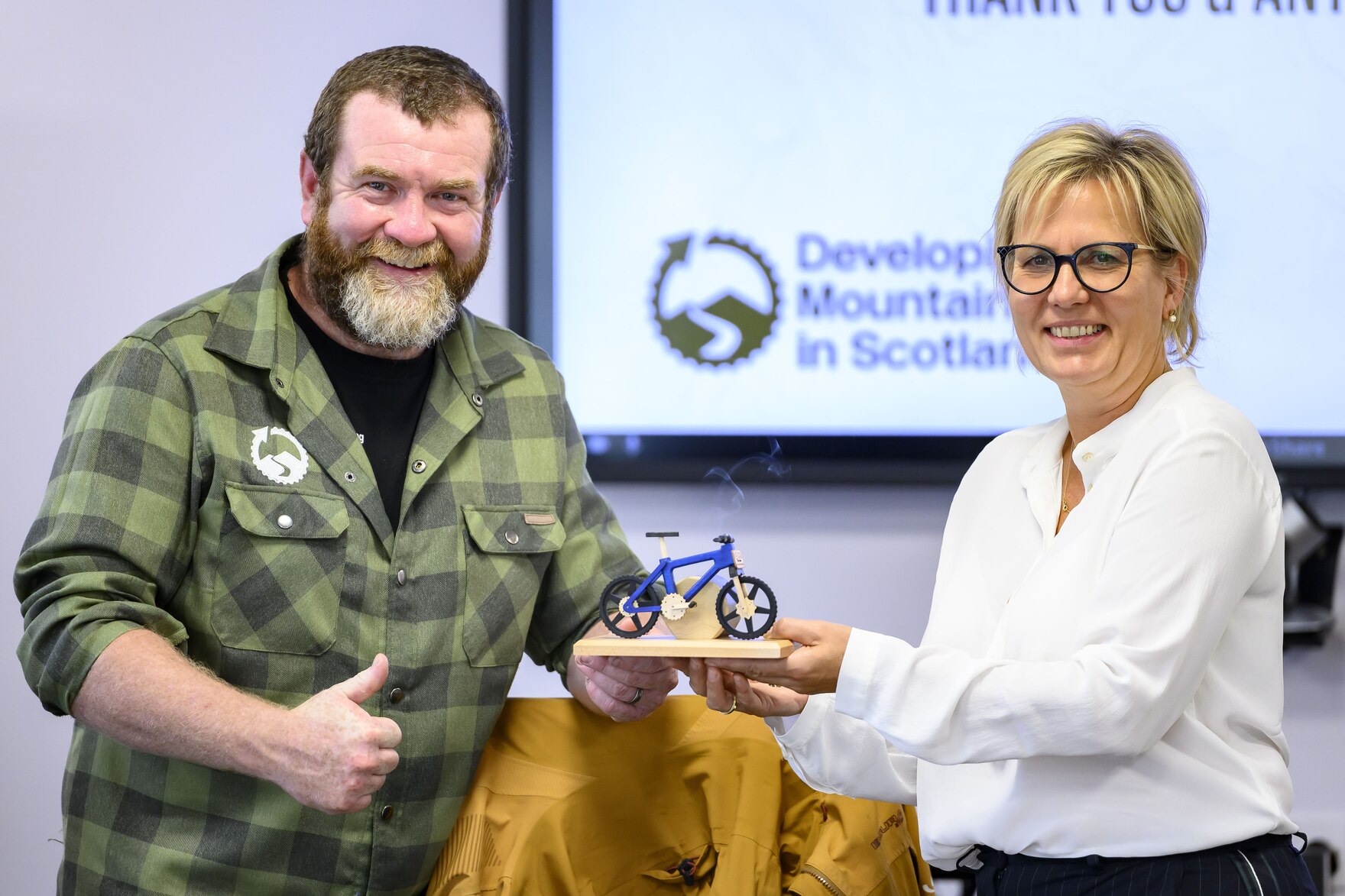 Die Ministerin übergibt dem schottischen Mountainbike-Experten ein Gastgeschenk: Ein Räuchermännchen in Mountainbike-Form